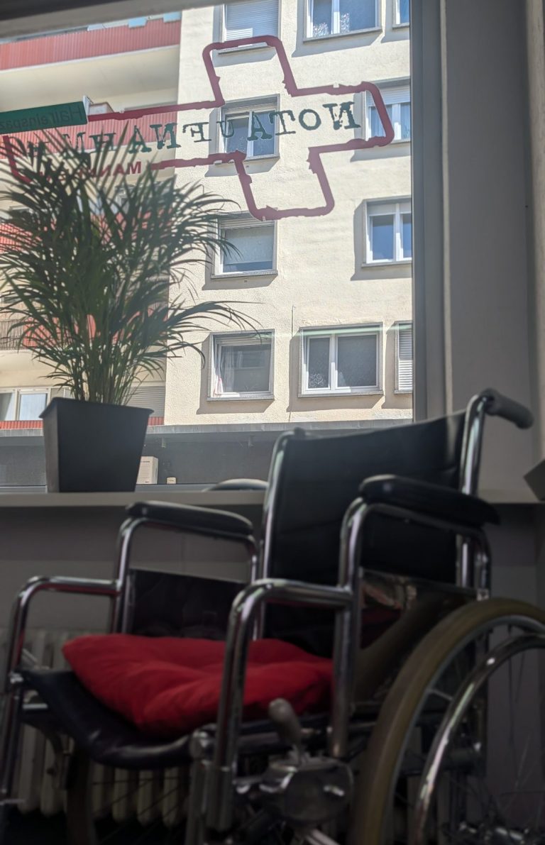 Rollstuhl Ein Rollstuhl mit rotem Kissen vor einem Fenster mit dem Notaufnahme Mannheim Logo und einer Pflanze.